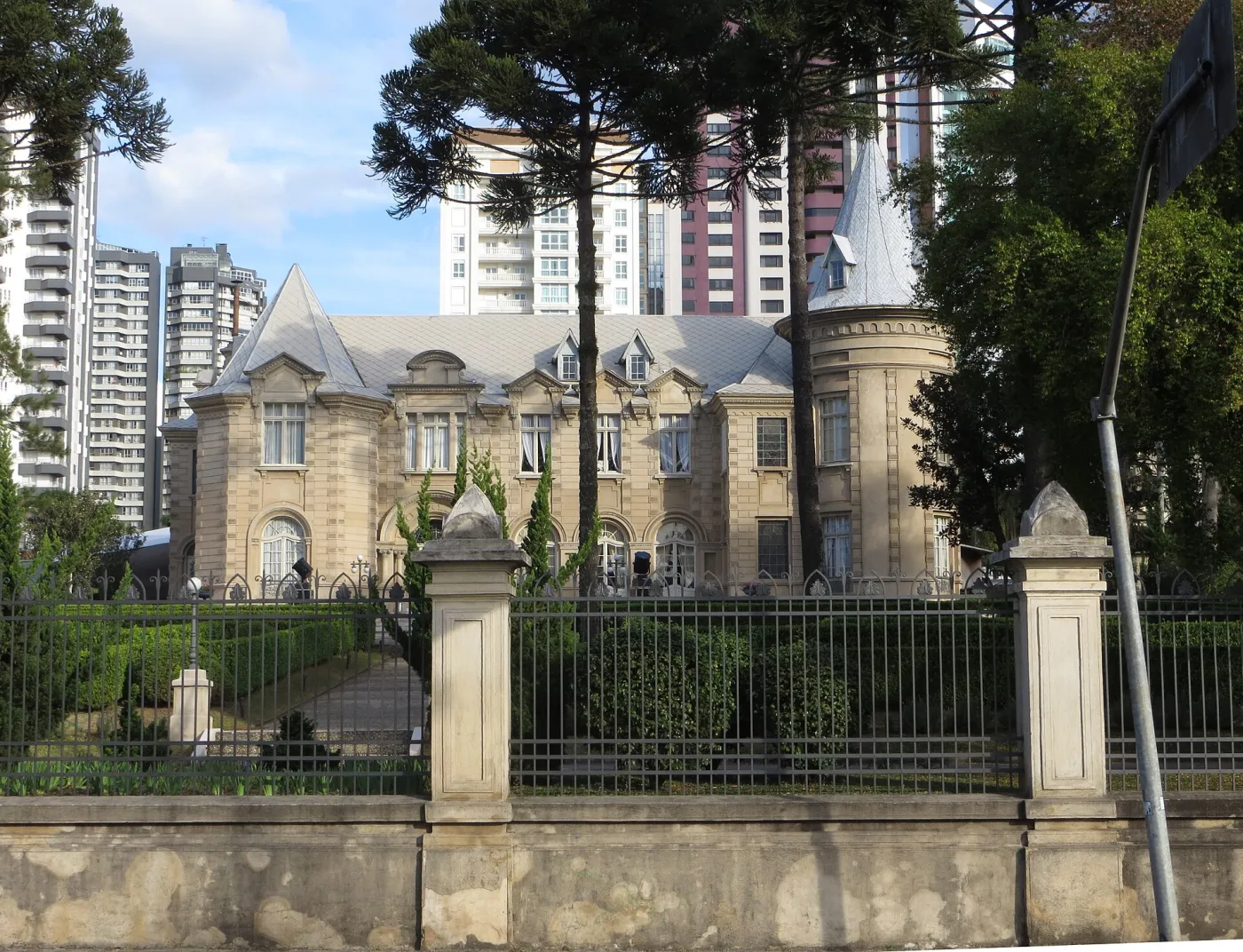 Portão e gradil de ferro do Castelo do Batel com pilares de pedra e araucárias ao fundo